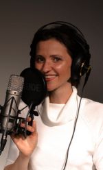 Frau mit Kopfhörern steht vor einem Mikrofon und lächelt. Sie trägt ein weißes Oberteil und spricht oder singt in ein Studiomikrofon